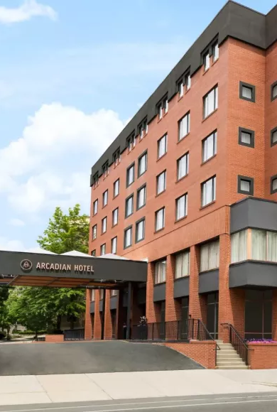 A brick midrise hotel building with a covered driveway, labeled &ldquo;Arcadian Hotel,&rdquo; set on a tree-lined street with clear blue sky.