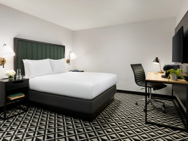 A modern hotel room with a king bed, green headboard, pair of wall lamps, a desk with chair, a flat-screen TV, and a black-and-white geometric patterned carpet.