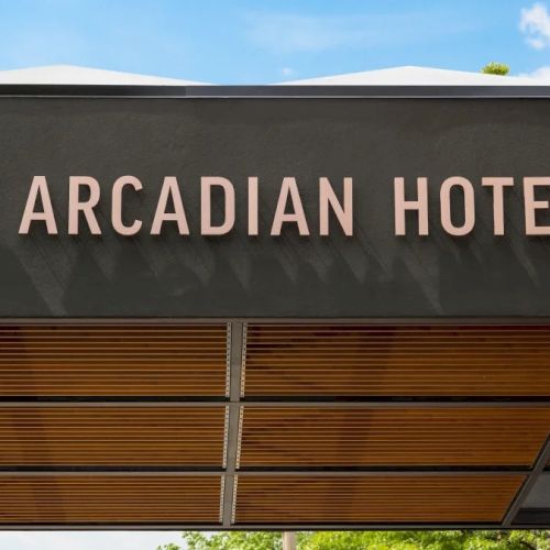 A sign reading &ldquo;Arcadian Hotel&rdquo; on a modern entrance canopy, with circular logo, metal letters, and a blue sky backdrop ending this line.