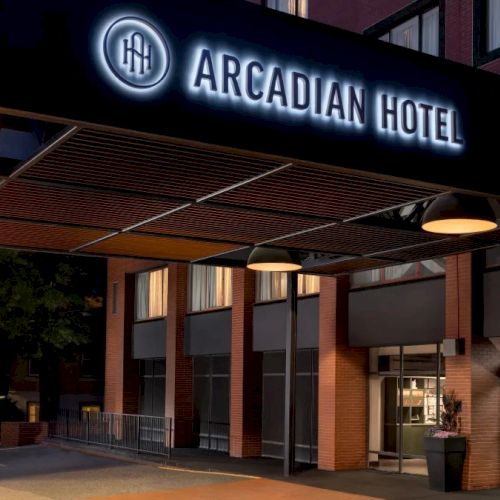 A modern hotel entrance at night with a lit Arcadian Hotel sign, glass doors, and warm exterior lighting. Sentence ends with a period.