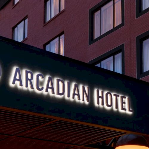 A neon sign reads &ldquo;Arcadian Hotel&rdquo; on a dark building facade with windows, suggesting a stylish hotel entrance at dusk.
