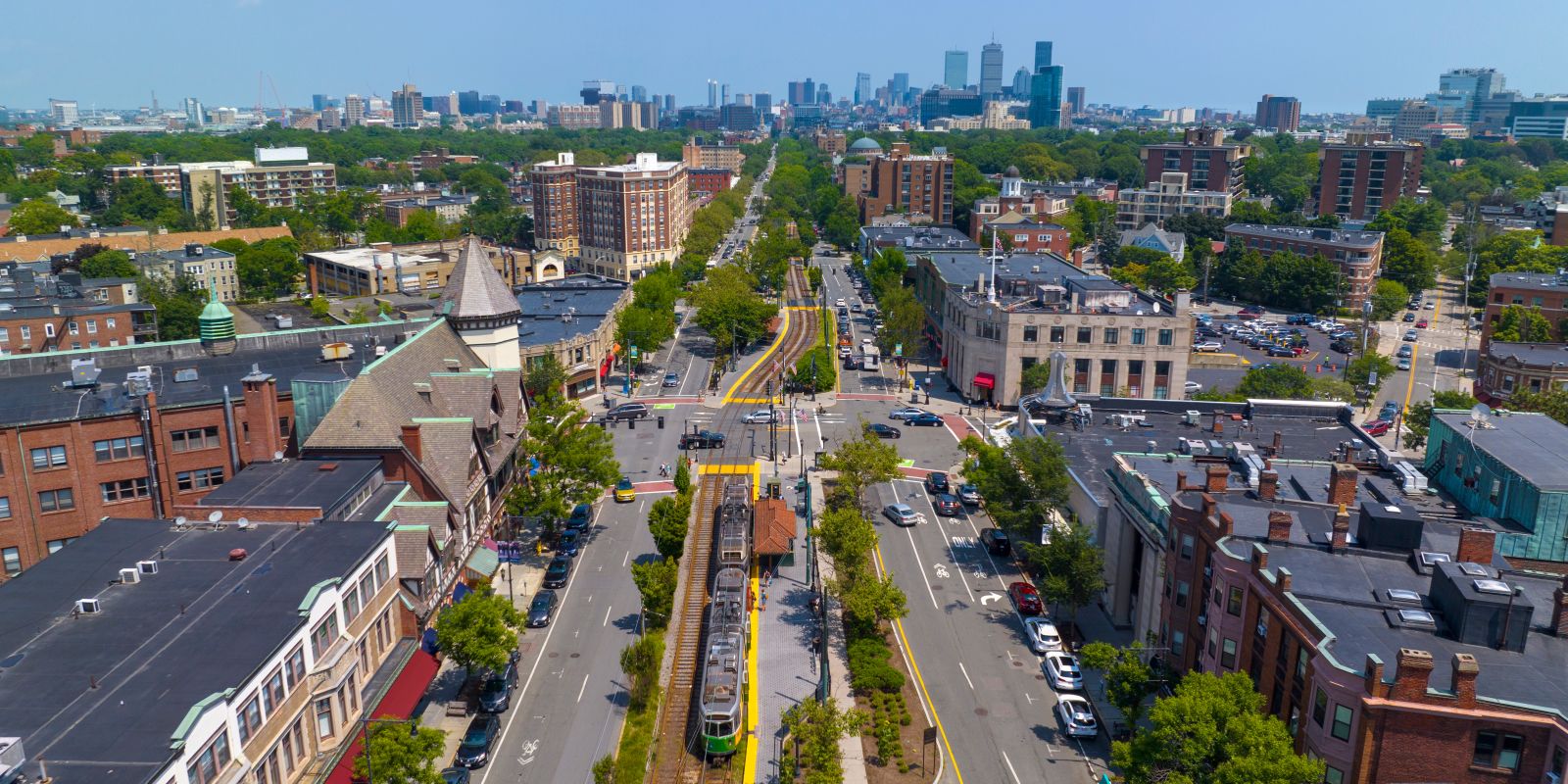 Aerial view of a city street lined with trees, buildings on both sides, a central tram line, and distant skyline on a clear day.