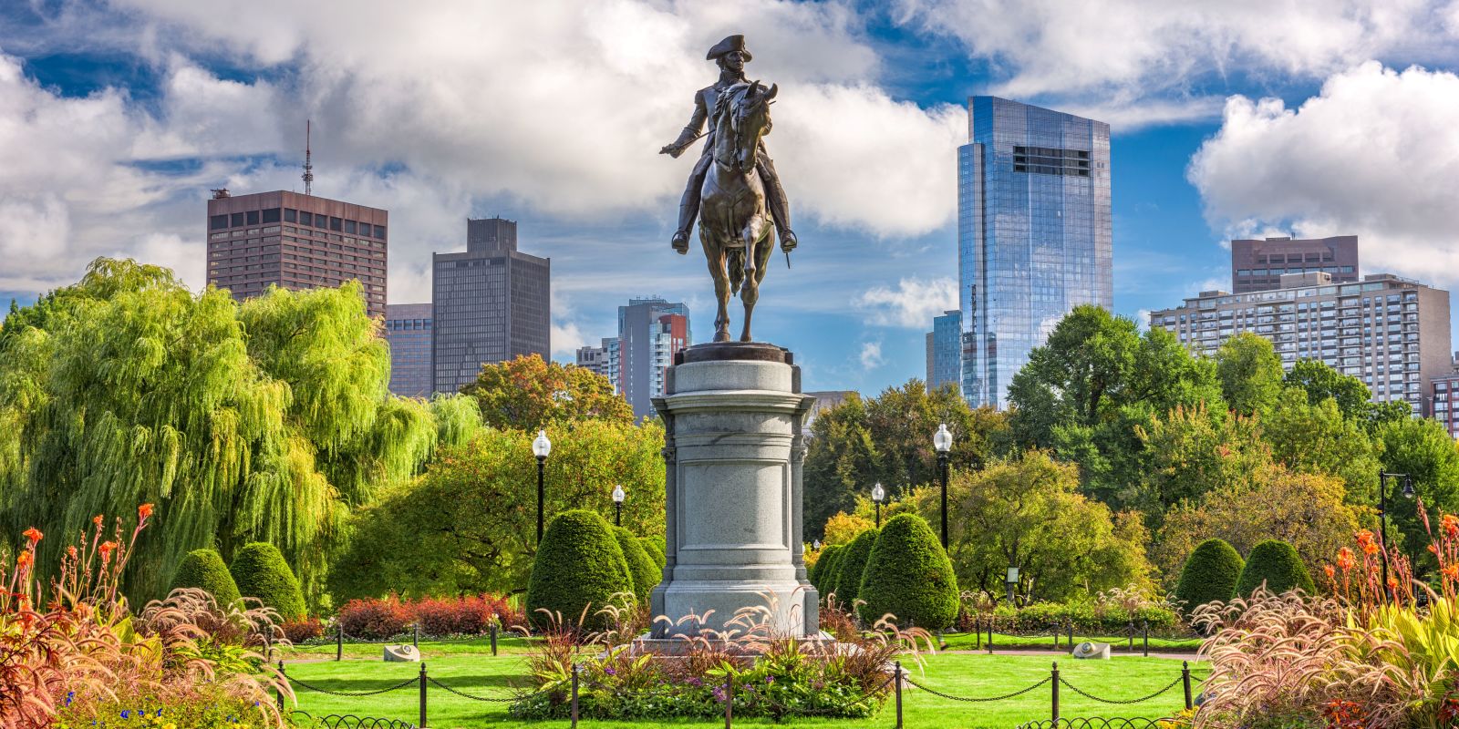 Statue of a rider on a tall pedestal in a park, with green trees, flowers, and a city skyline in the background under a blue, partly cloudy sky.