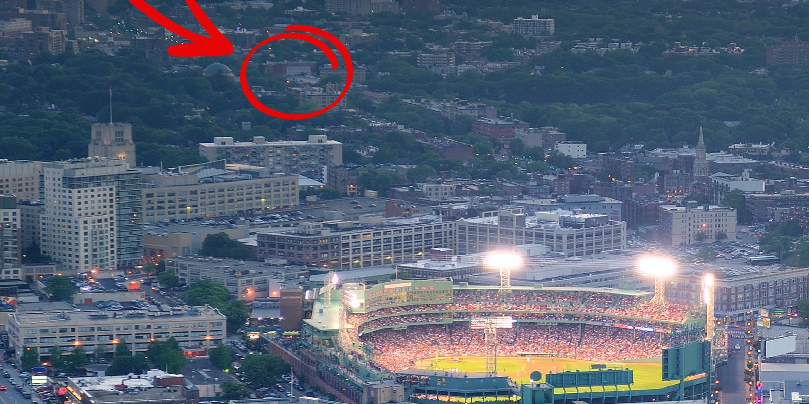 A cityscape with a stadium glowing at dusk; a distant circled area shows a faint tower or building behind trees, barely visible in the distance.